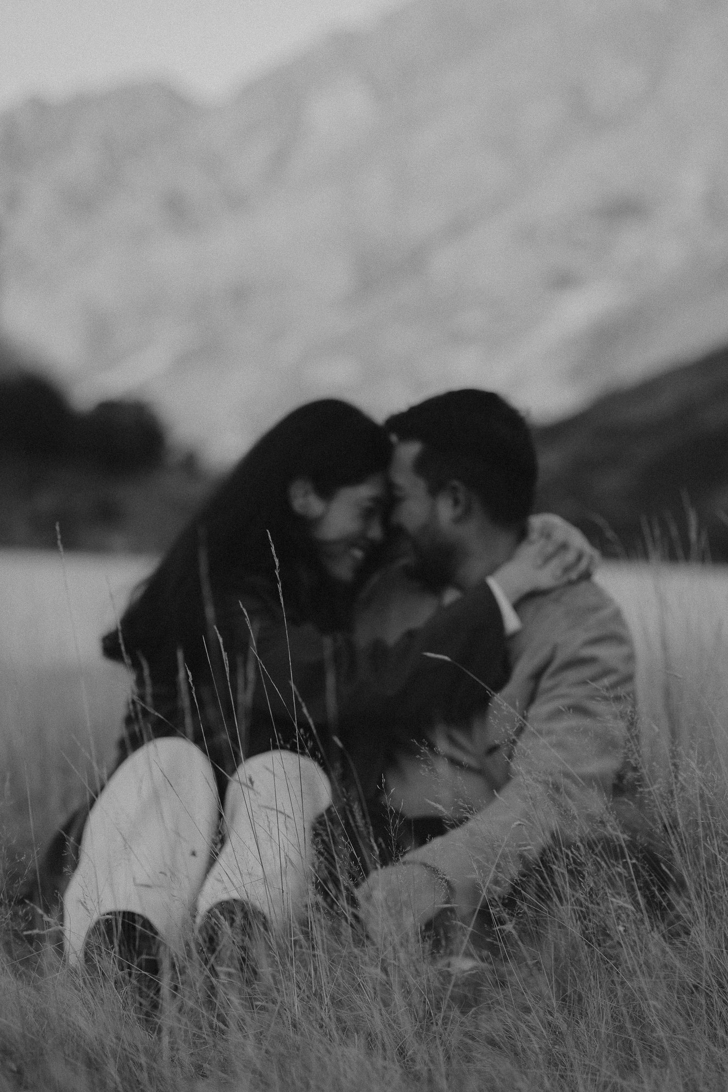 Black and white photo of a couple sitting in tall grass, sharing an intimate moment with mountains in the background.