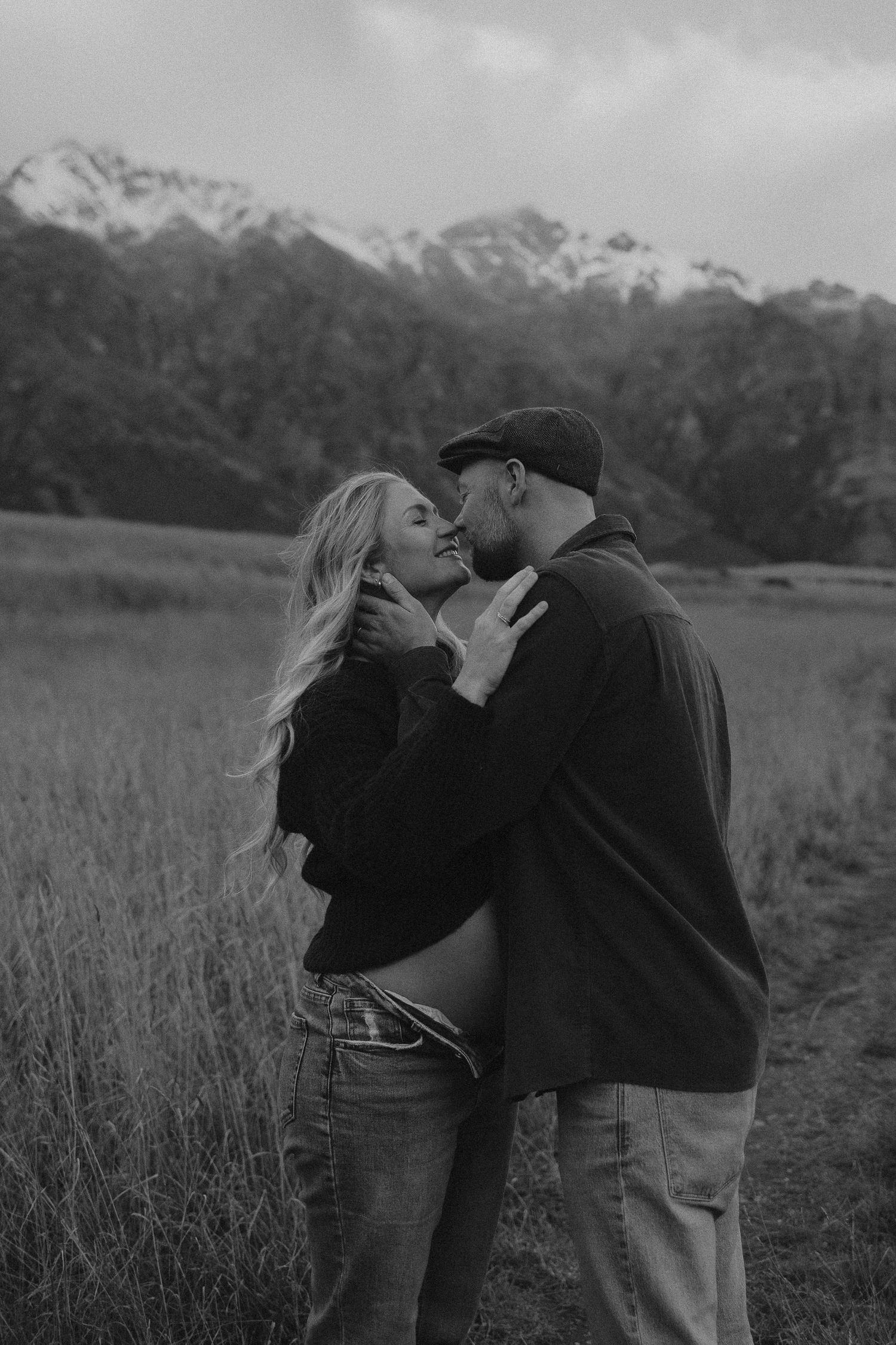 Black and white photo of a couple sharing a romantic moment in a field with snow-capped mountains in the background.