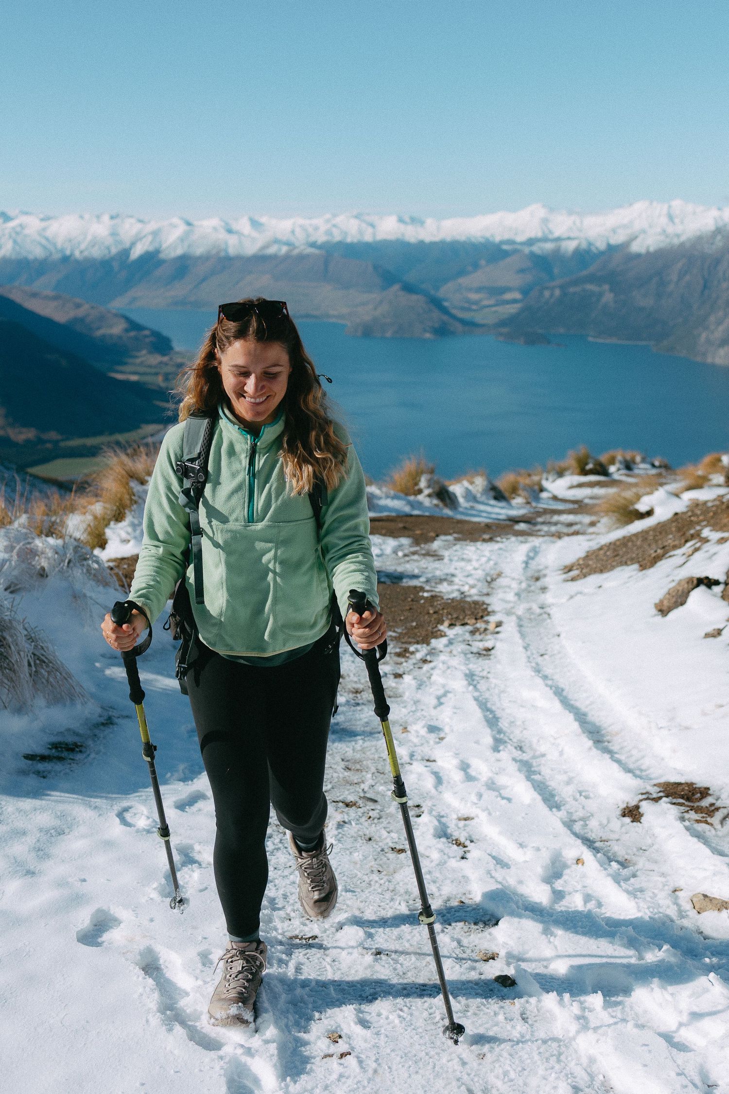 Woman hiking on snowy mountain trail with trekking poles, scenic lake and snow-capped mountains in background.