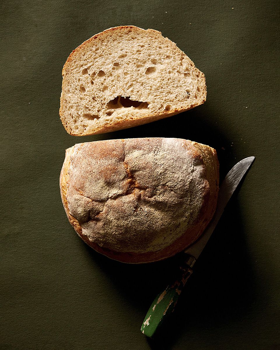 Rustic sourdough bread loaf sliced open on dark surface with a knife, showing airy crumb texture.
