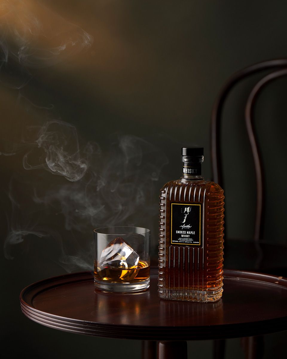 Bottle of whisky beside a glass with ice on a dark wooden table with dramatic smoky atmosphere.