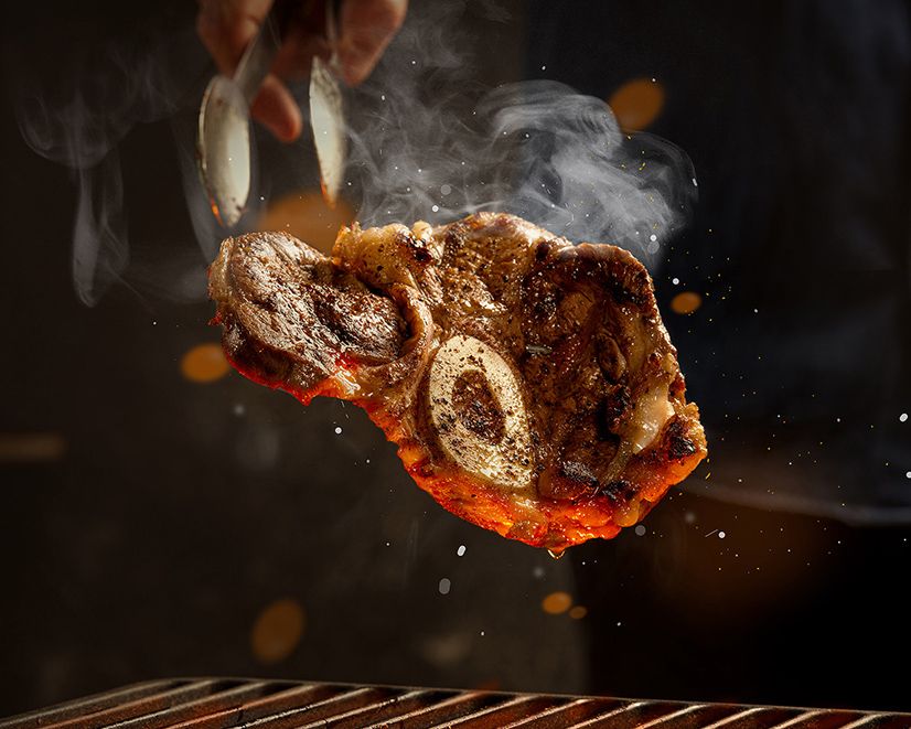 Chef grilling a juicy bone-in steak with tongs over hot grill grates, steam rising dramatically in dark kitchen.