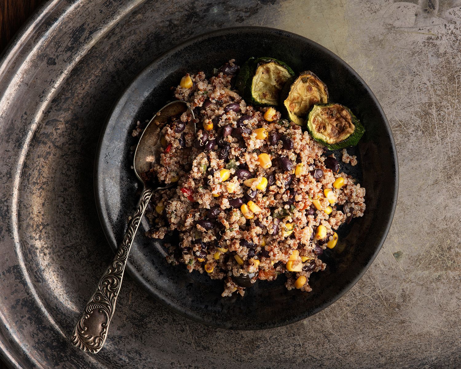 Black bowl of quinoa salad with corn, black beans, and roasted Brussels sprouts on a rustic metal tray.