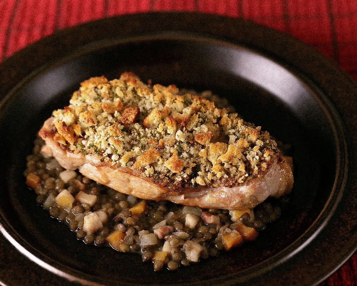 Herb-crusted pork chop served on a bed of seasoned lentils in a dark bowl on a red surface.