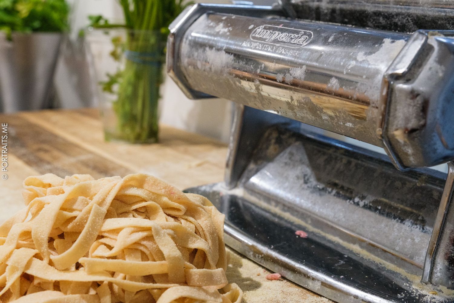 Fresh homemade fettuccine pasta beside a vintage Imperia pasta machine on a flour-dusted wooden surface.