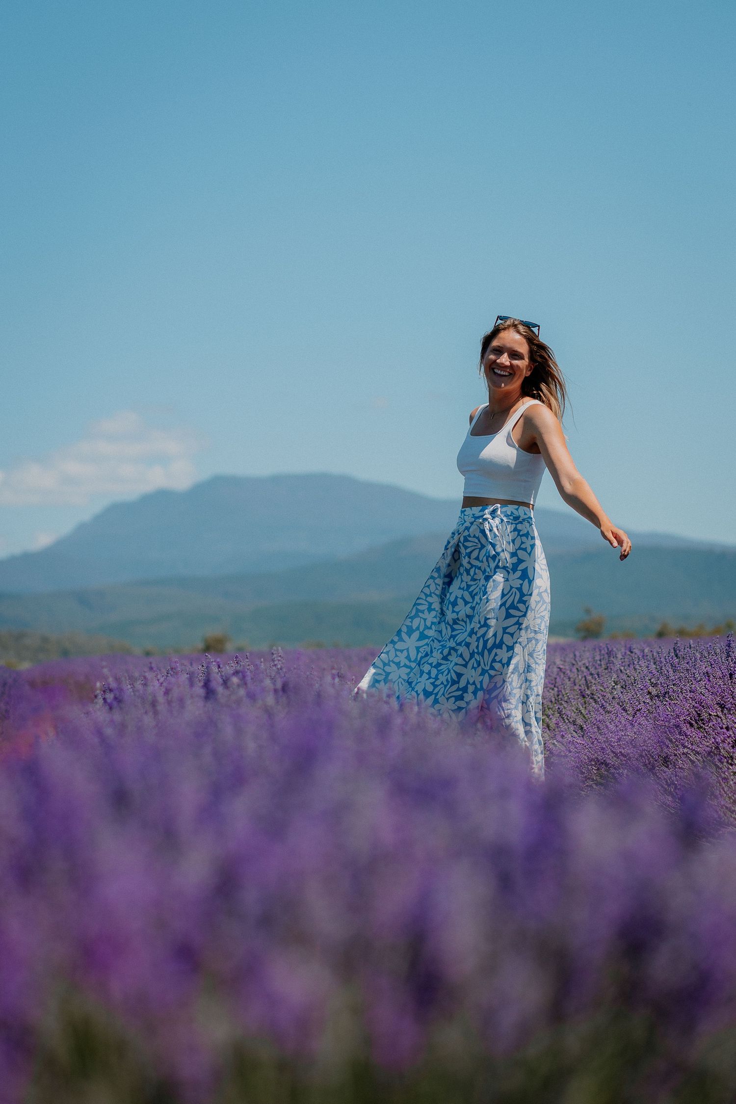 Woman in white crop top and blue floral skirt smiling in a vibrant purple lavender field with mountains in background.