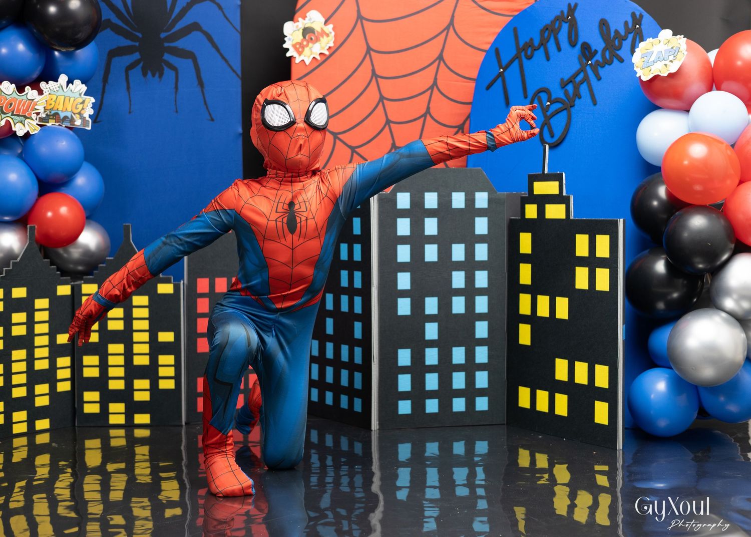 Person in Spider-Man costume poses at a birthday party with cityscape backdrop, colorful balloons, and Happy Birthday sign.