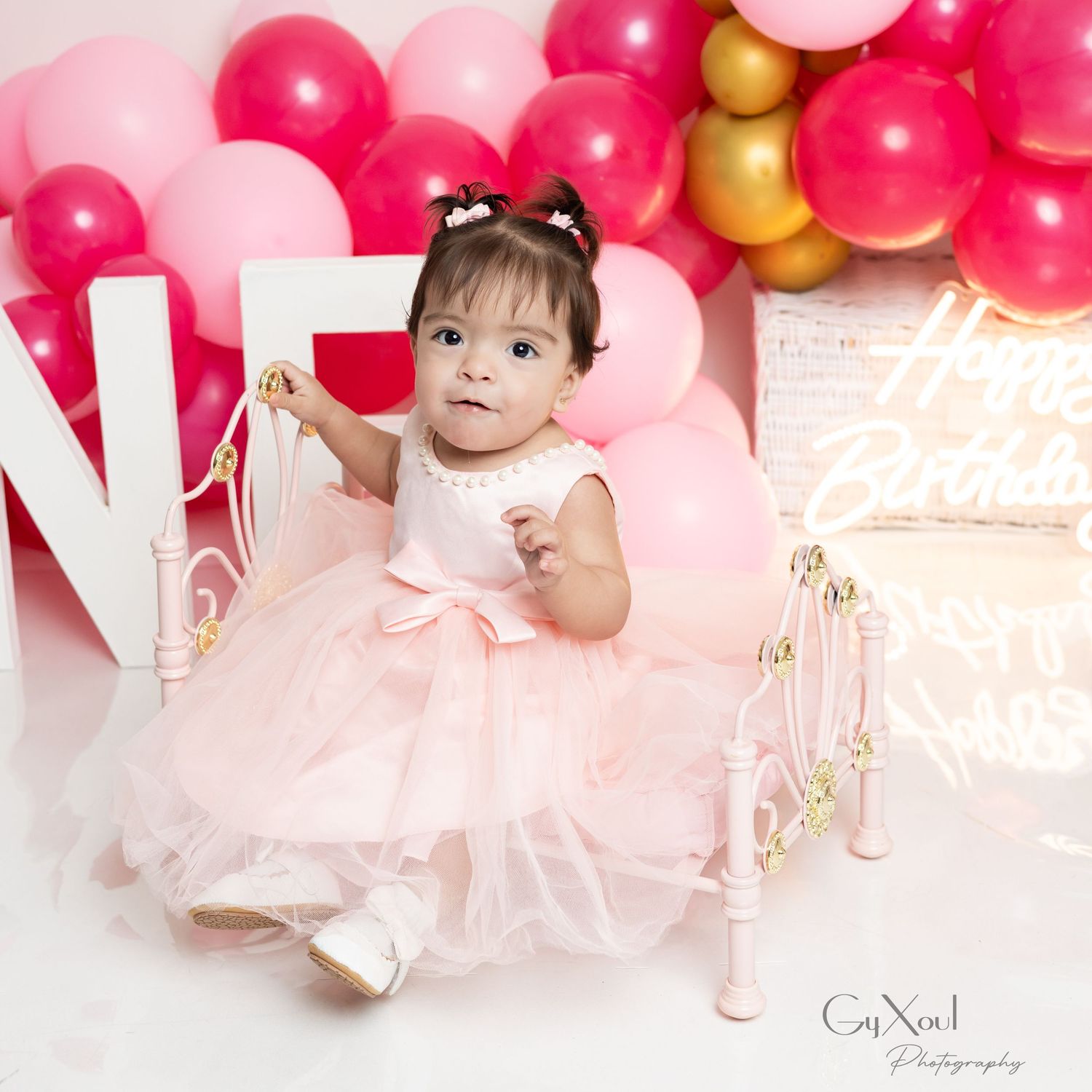 Baby girl in pink tutu dress sits for first birthday cake smash photo with balloons and Happy Birthday neon sign.