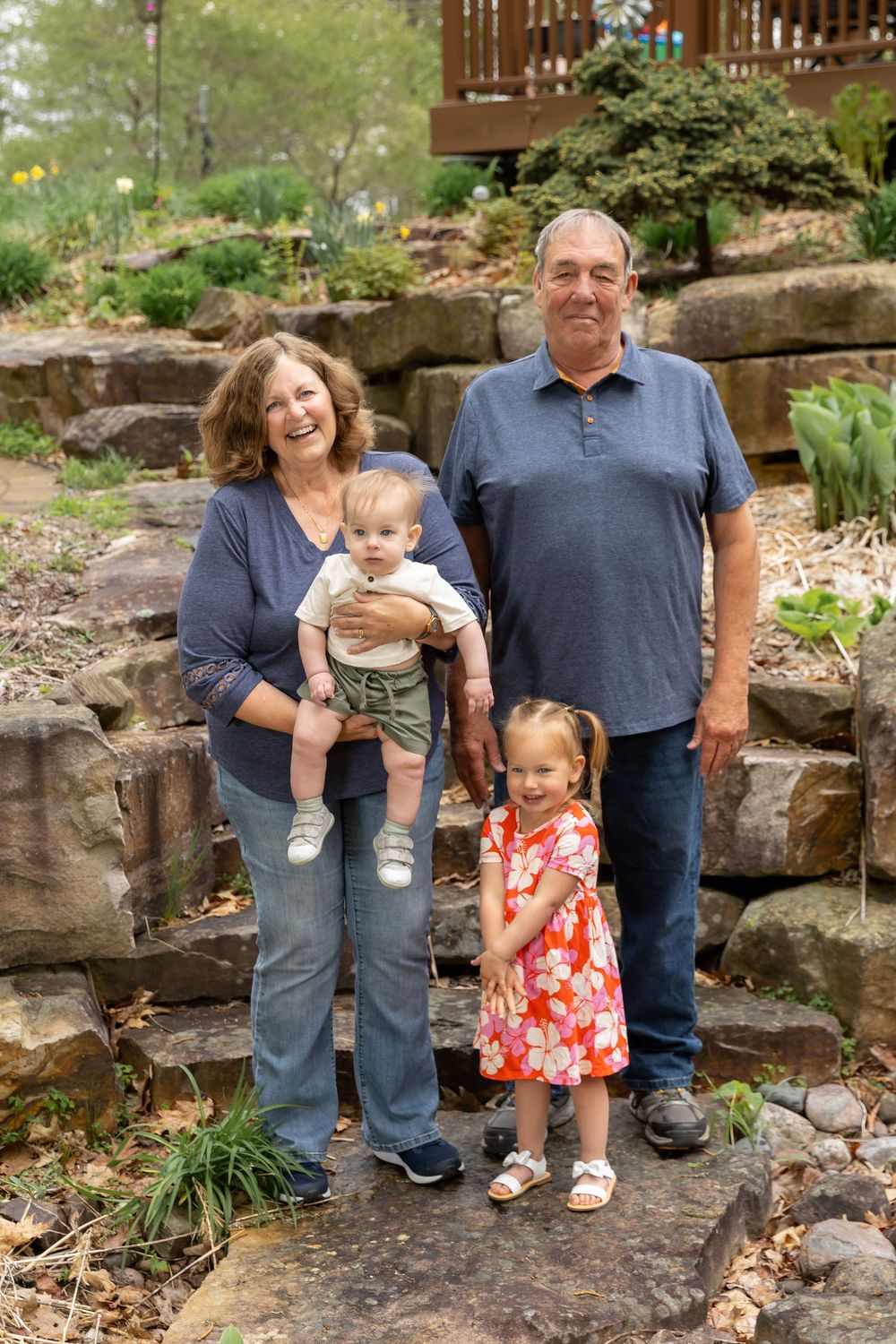 Grandparents posing outdoors with two young grandchildren on stone steps surrounded by a lush garden.
