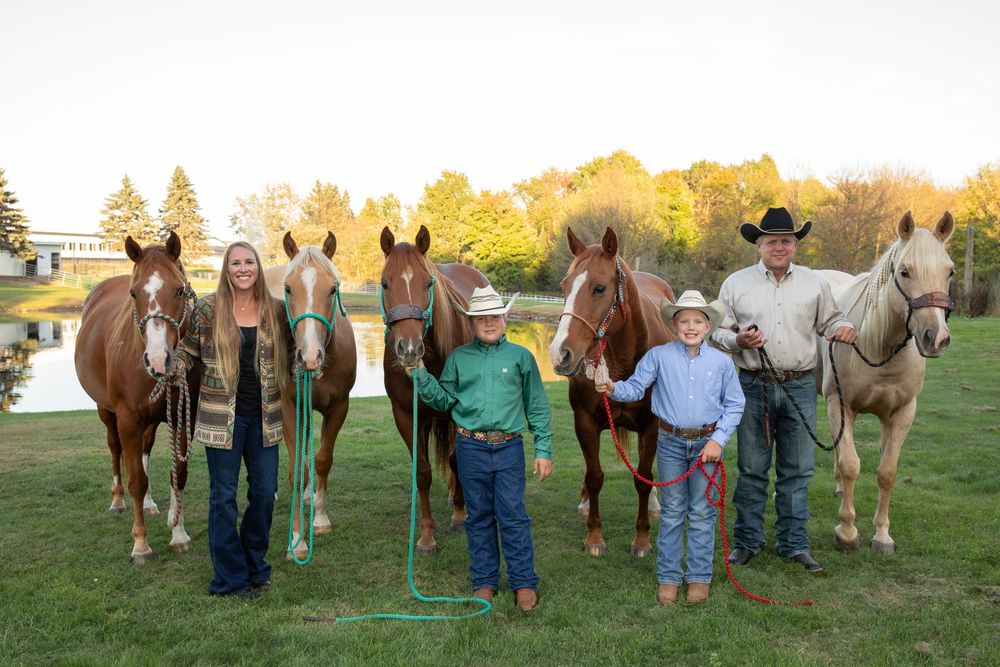 Family of four posing outdoors with five horses on green grass at golden hour near a pond.