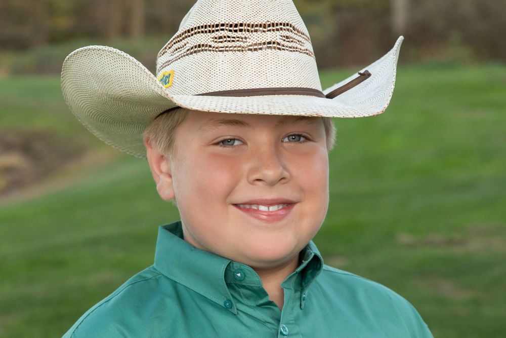 Smiling young boy wearing a white cowboy hat and green shirt outdoors.