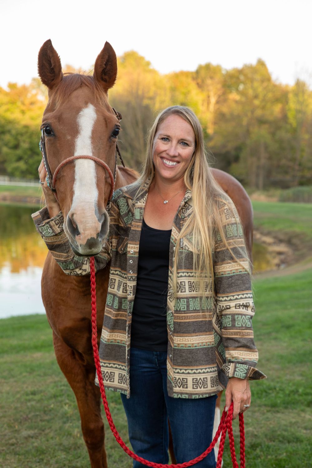 Smiling blonde woman standing with a chestnut horse with a blaze, holding a red lead rope near a pond.