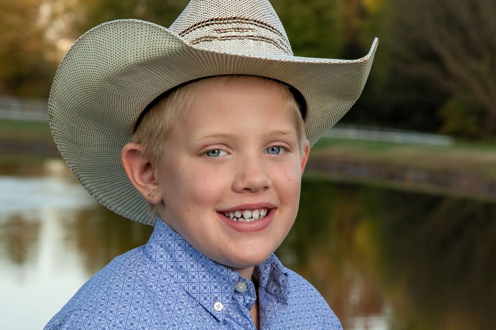 Smiling blonde boy wearing a cowboy hat and blue shirt outdoors near a pond.