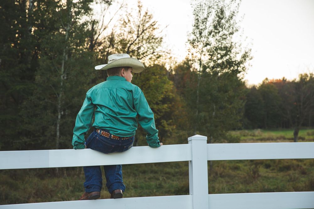 A cowboy in a green shirt and hat leans on a white fence overlooking a green pasture.