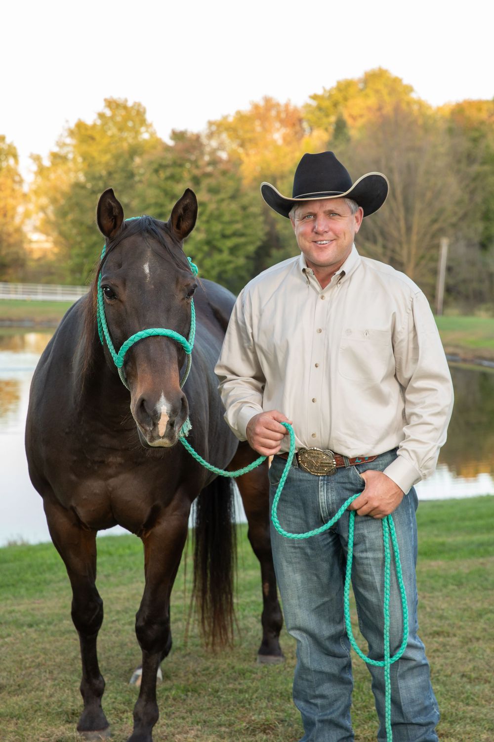 Man in black cowboy hat holding teal rope lead with dark bay horse outdoors near pond.