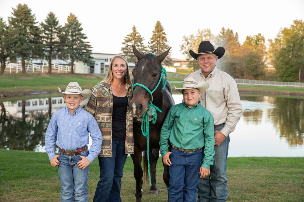 Family of four in western wear posing with a dark horse by a pond on a sunny day.