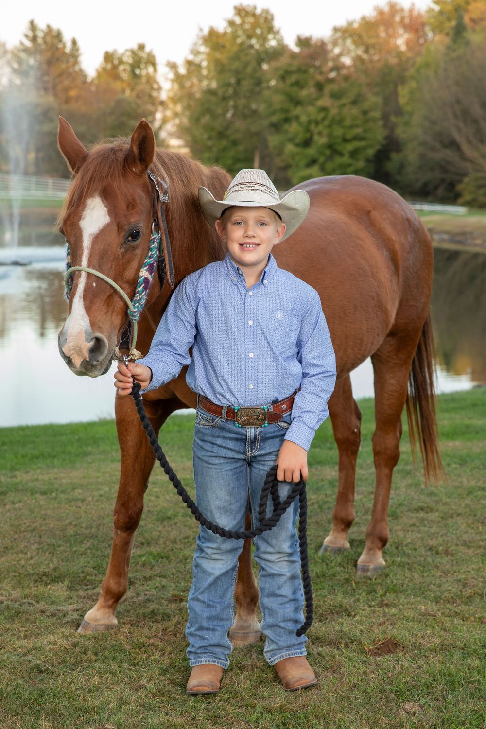 Young boy in cowboy hat smiling beside chestnut horse holding black lead rope outdoors.