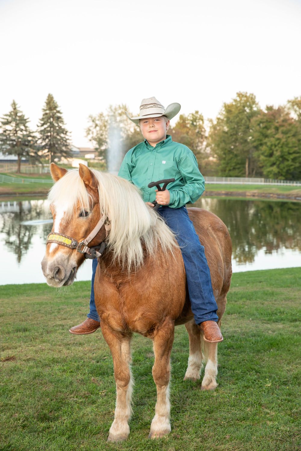 Young boy in cowboy hat and green shirt riding a palomino pony near a pond outdoors.