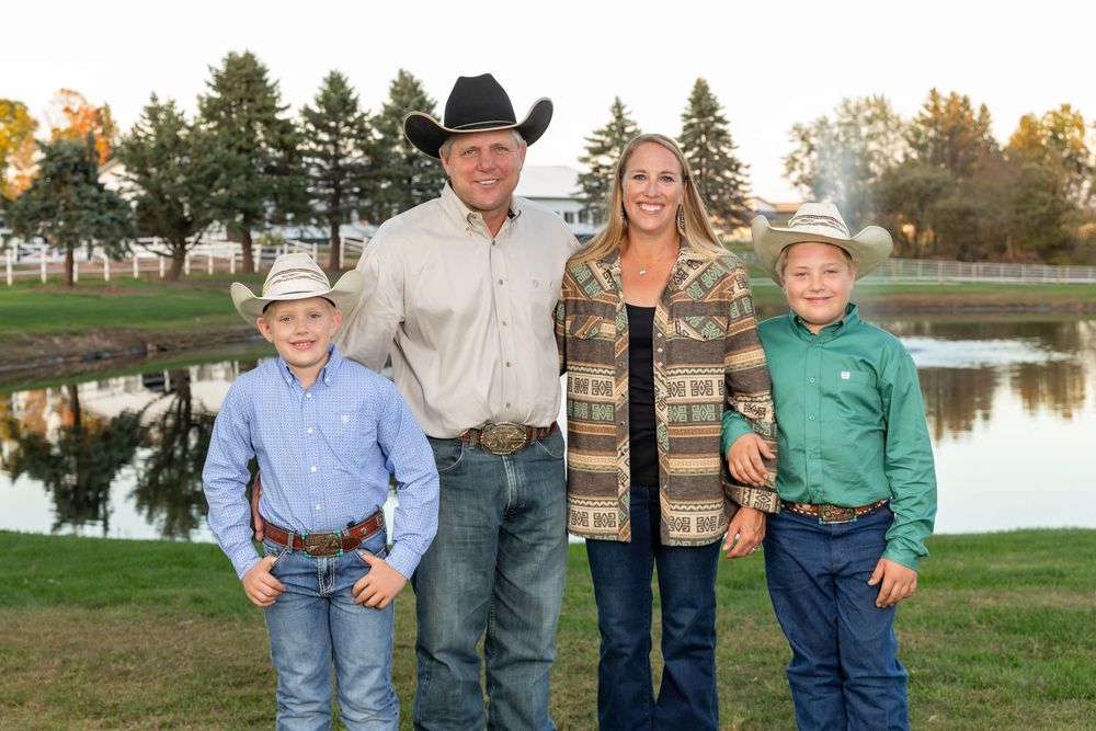 Family of four in western wear smiling outdoors near a pond on a sunny autumn day.