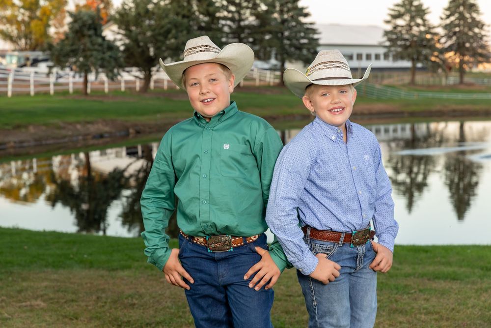 Two smiling boys in cowboy hats and western shirts posing together near a pond outdoors.