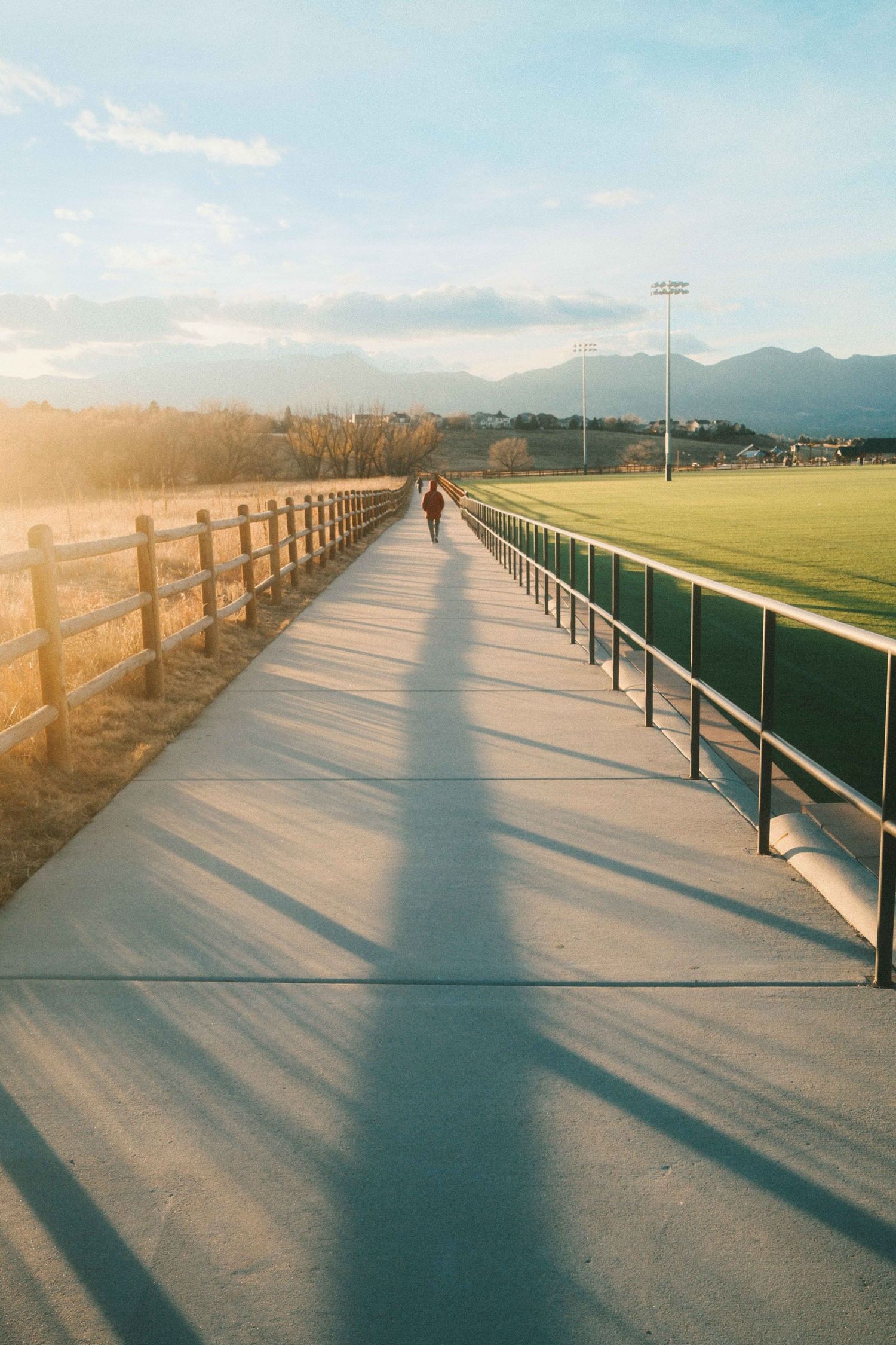 A person walks along a sunlit concrete bridge path beside a green sports field with mountains in the background.