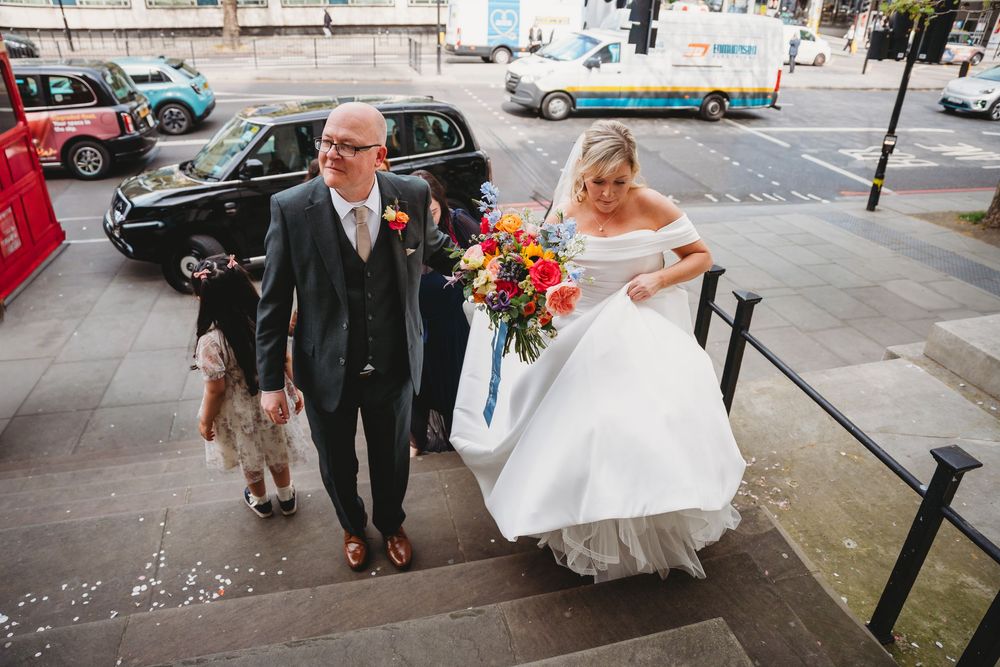 Bride in white gown holding colorful bouquet walks with her dad in green suit walking up the stairs of Marley-bone town hall