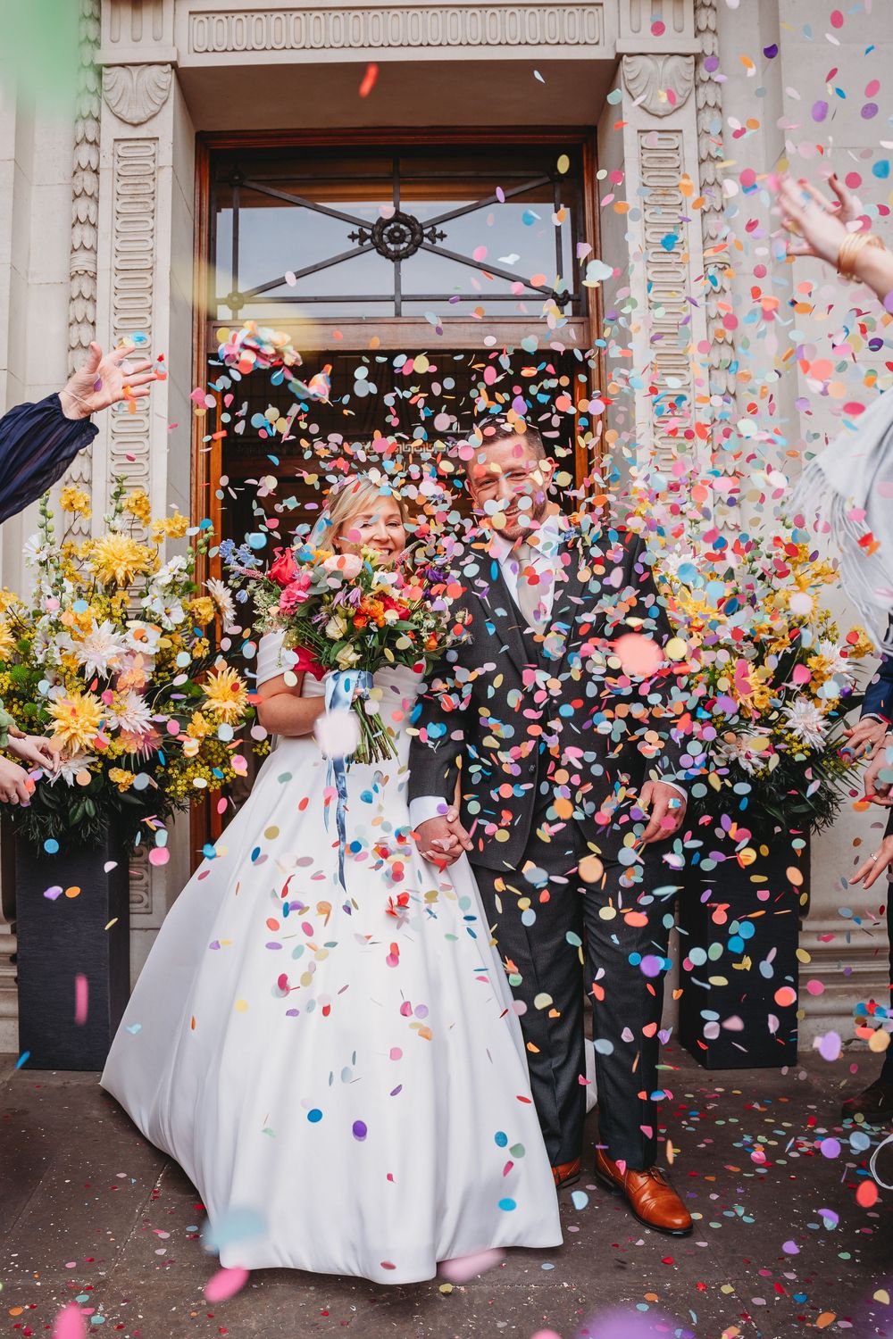 Joyful newlyweds exit venue doors as guests shower them with colorful confetti outdoors.