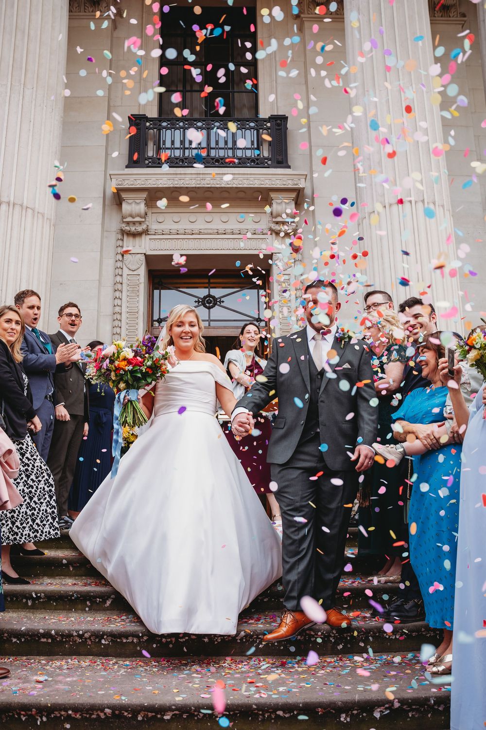 Newlyweds exit grand building steps amid colorful confetti shower, surrounded by cheering wedding guests.