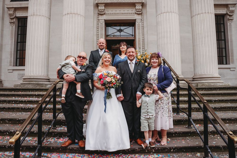Wedding party poses on stone steps outside a grand building with colorful confetti scattered around.