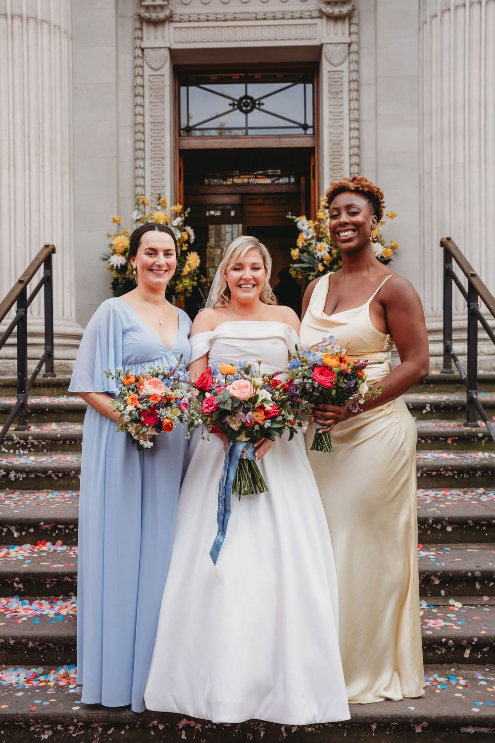 Bride with two bridesmaids in blue and gold dresses holding colorful bouquets on confetti-covered stone steps.