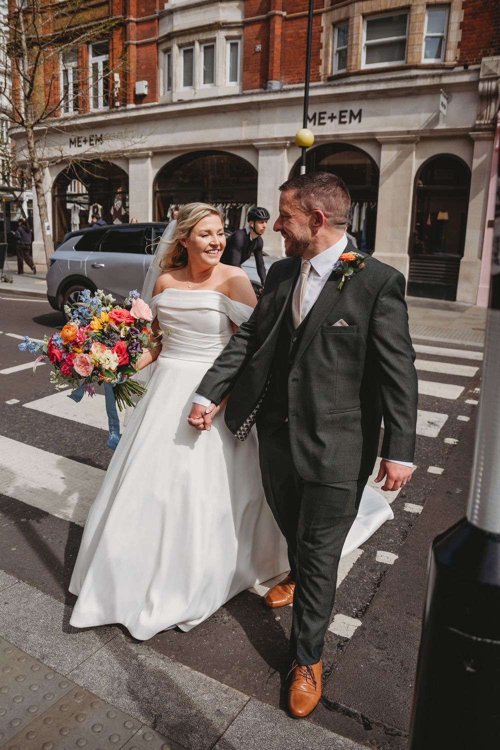 Bride and groom holding hands crossing a London street, bride holding colorful bouquet in white off-shoulder gown.
