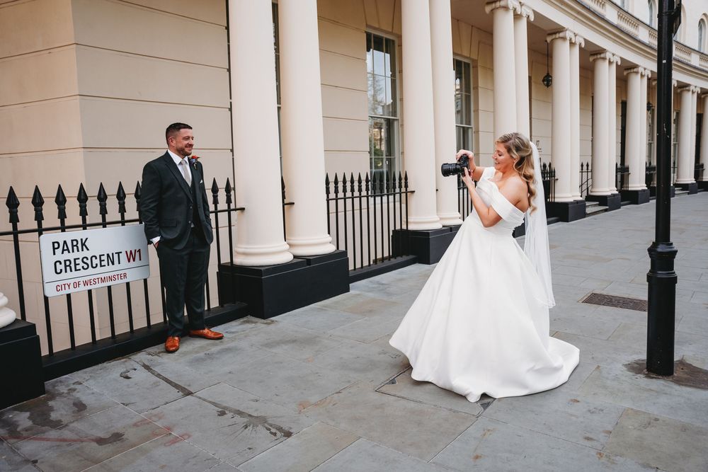 Bride in white ball gown photographs groom outside Park Crescent West building with white columns.