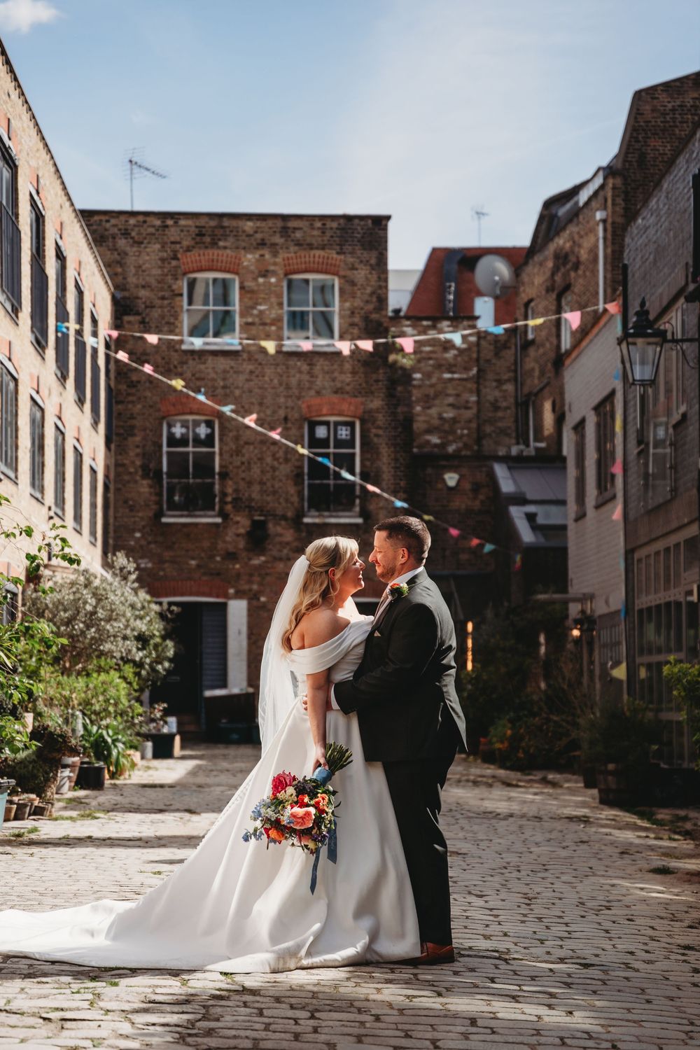 Bride and groom share a romantic moment on cobblestone street with brick buildings.