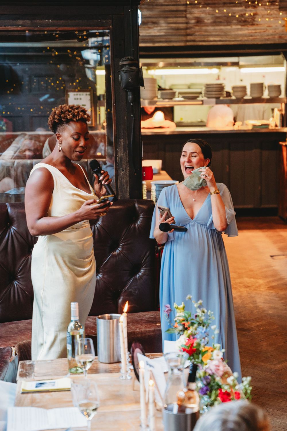 Two bridesmaids giving emotional speech at wedding reception in rustic restaurant venue.