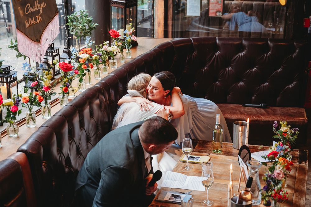 Bride embraces a guest warmly at a decorated reception table with colorful floral centerpieces.