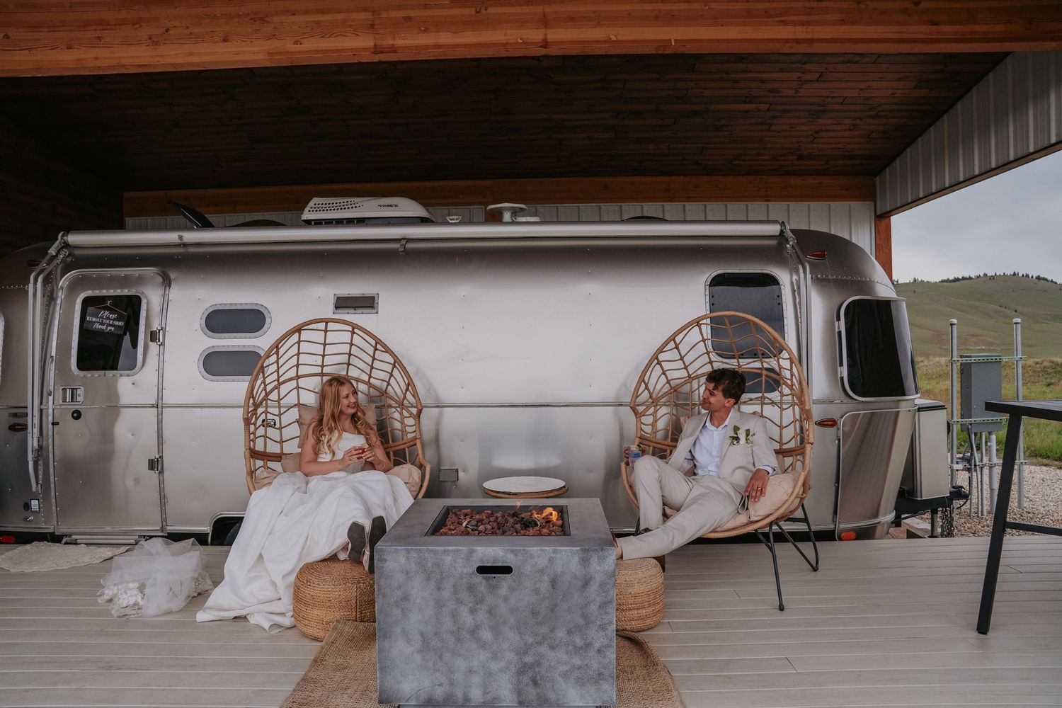 Bride and groom relaxing in wicker chairs beside an Airstream trailer with a fire pit on a covered patio.