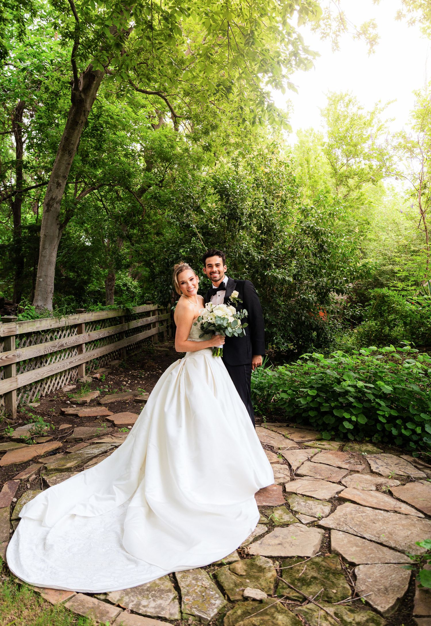Bride in white gown with train and groom in black tuxedo pose on stone path surrounded by lush green garden trees.