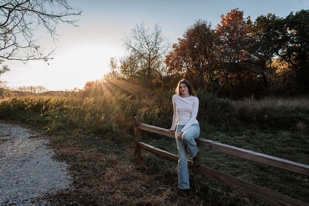Young woman in white sweater and jeans sits on a wooden fence at golden hour with autumn trees in background.