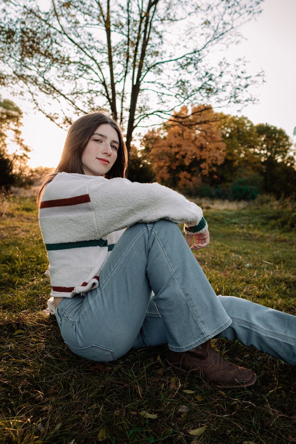 Young woman in striped sweater and jeans sitting on grass in autumn park at golden hour sunset.