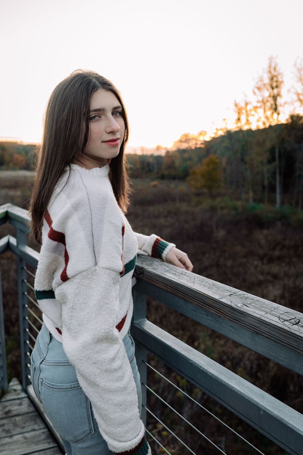 Young woman in white sweater and jeans leaning on wooden bridge railing at sunset with autumn trees in background.