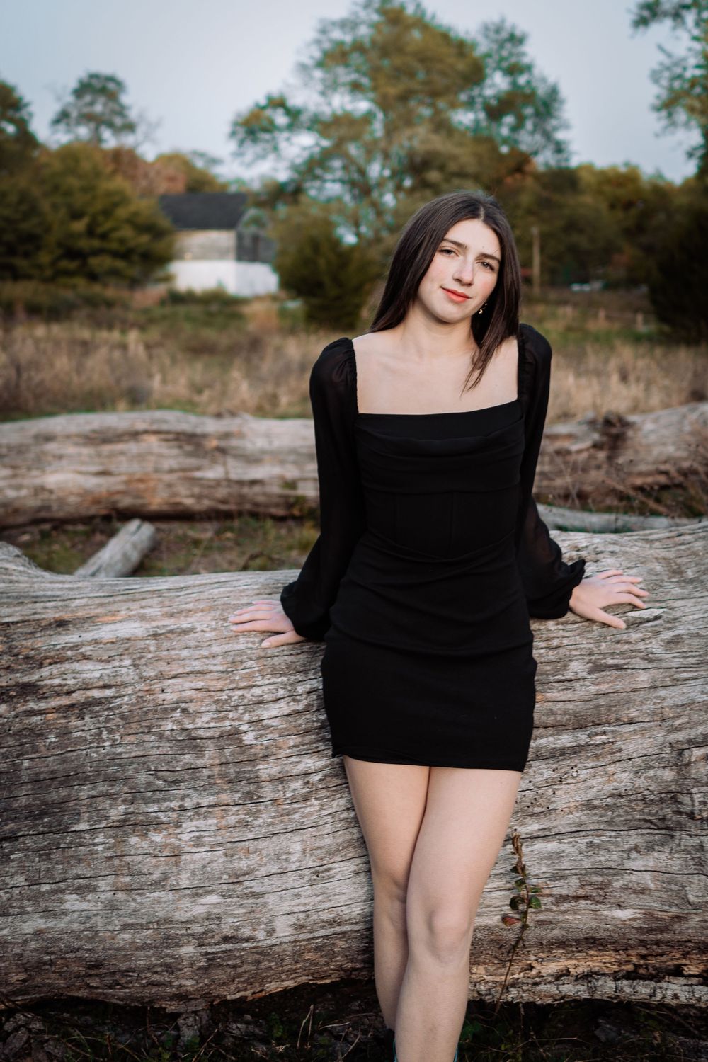 Young woman in black long-sleeve mini dress poses against a fallen log in a natural outdoor setting.