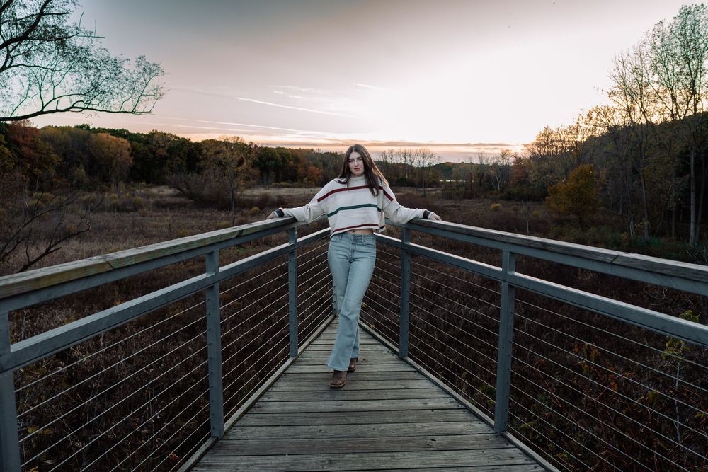 Young woman poses on a wooden bridge overlook at sunset, surrounded by autumn trees and a scenic landscape.