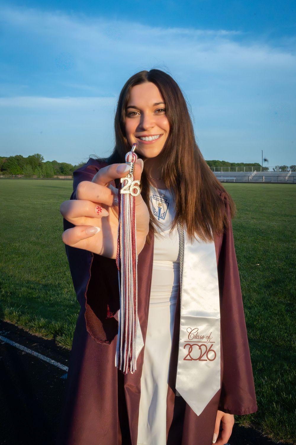 Smiling graduate in maroon cap and gown holds Class of 2026 tassel outdoors on a sunny day.