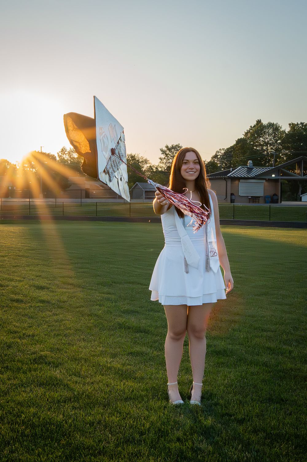 Young woman in white dress holding pom-poms and a sign on a green field at sunset during graduation.
