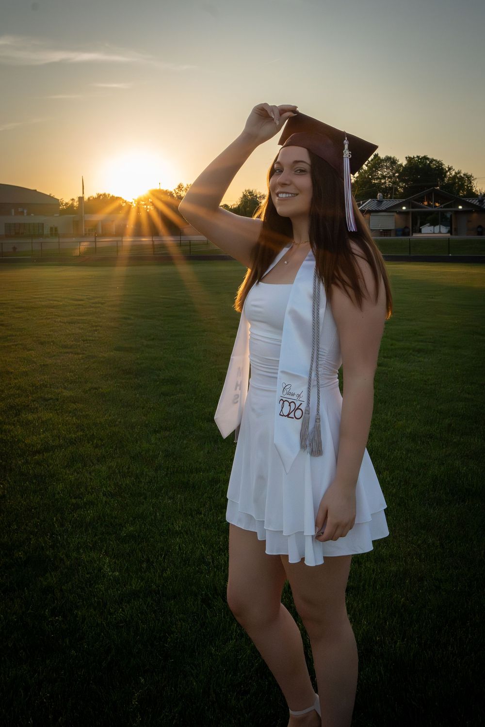Young woman in white dress and graduation cap smiling at sunset on green field.