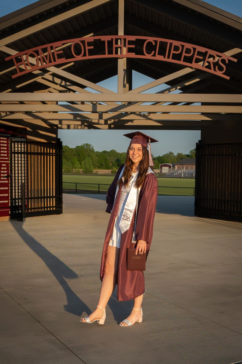 A smiling graduate in maroon cap and gown poses under a "Home of the Clippers" sign at golden hour.