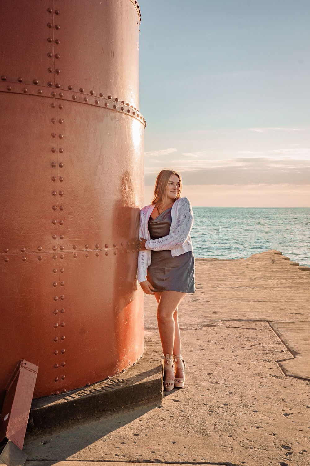 Young woman in white cardigan and gray skirt leaning against a red riveted structure by the ocean at sunset.