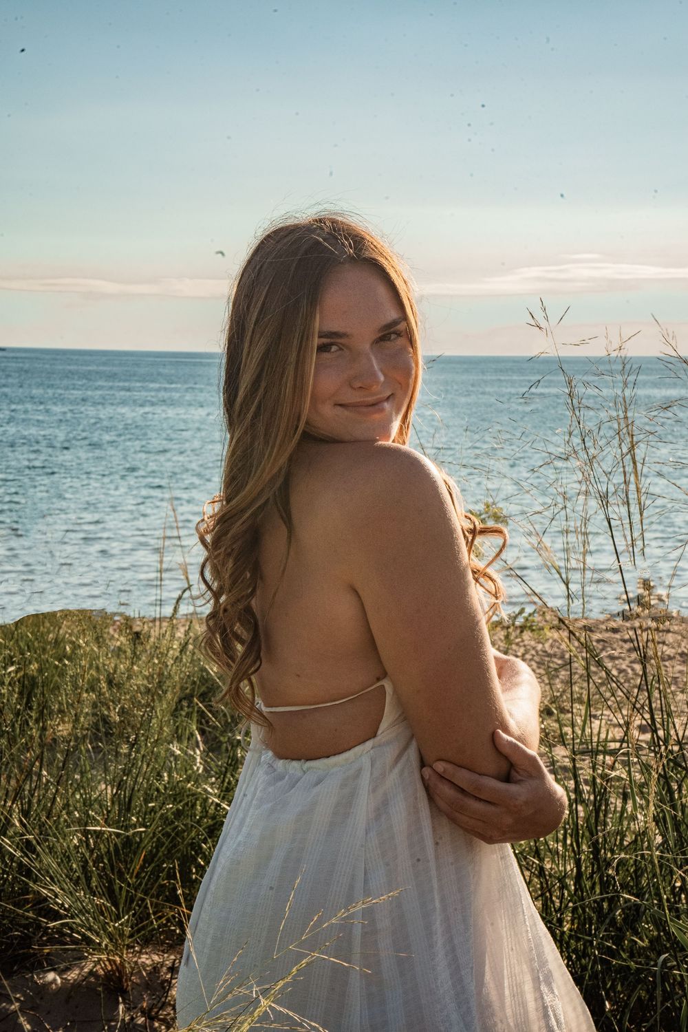 Young woman in white backless dress smiling over shoulder near ocean shore with tall grass at golden hour.