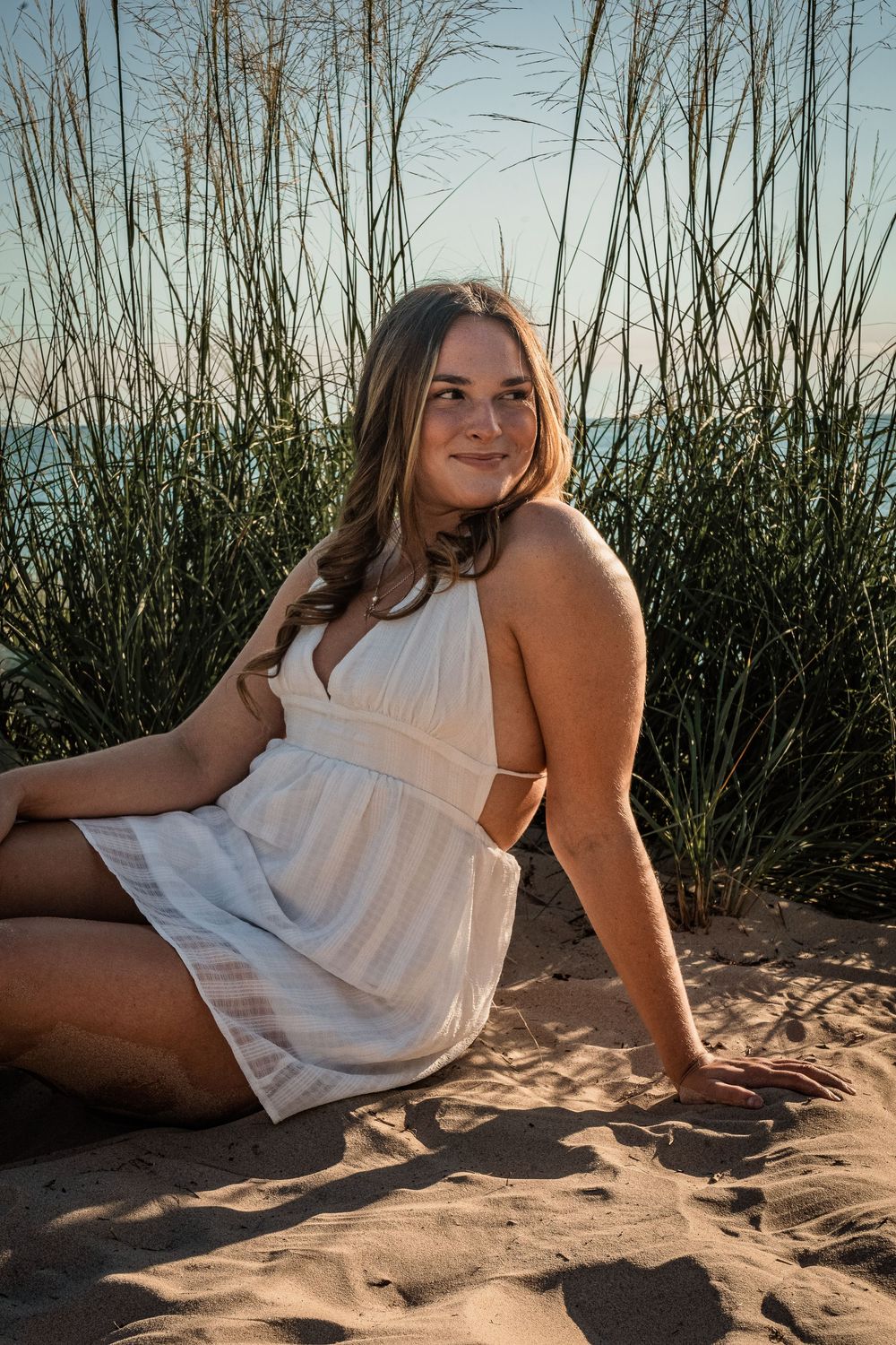 Young woman in white sundress sitting on sandy beach dunes with tall grass and ocean in background.