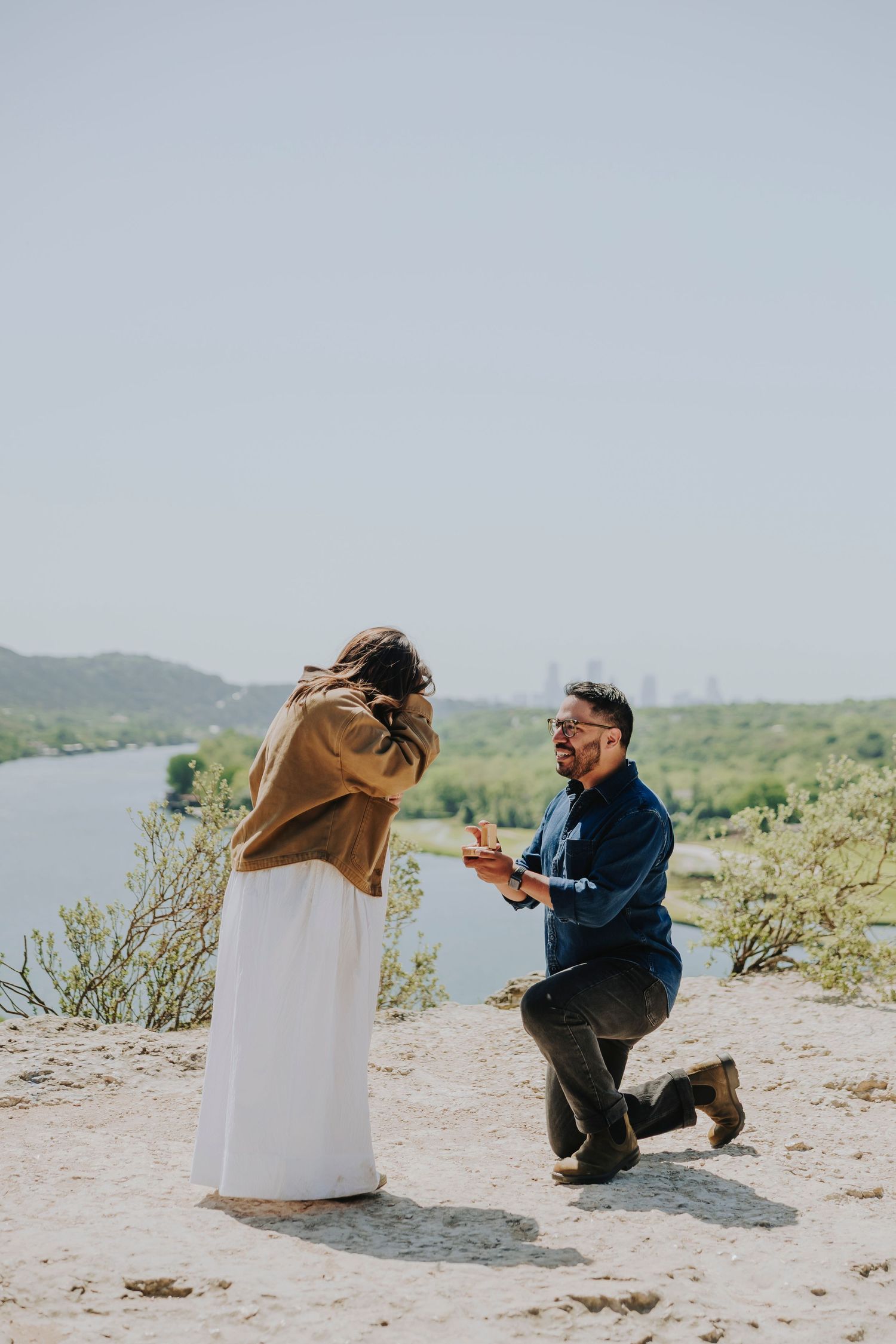 Man proposes on one knee to woman outdoors, holding ring box at rocky scenic location during golden hour.